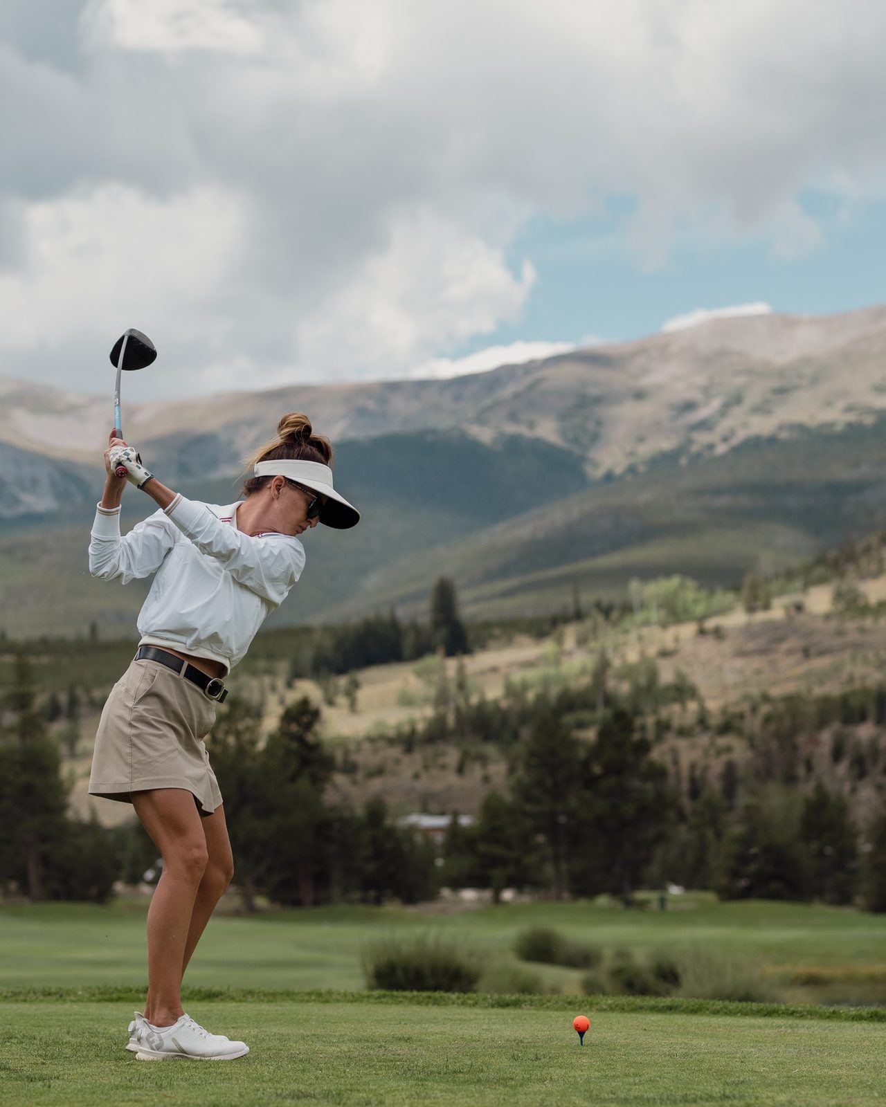 A woman at the top of her backswing on a Breckenridge tee, with the Colorado peaks behind her