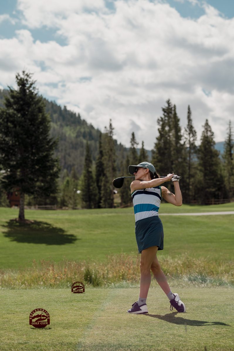 A player finishing her drive off the tee, with pines and mountains behind her